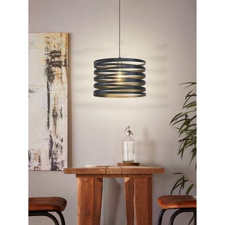 Pranvorne Pendant Light 37cm in black, a drum-shaped fixture of spiralling flat steel bands on a slim black cord, shown lit above a wooden dining table.