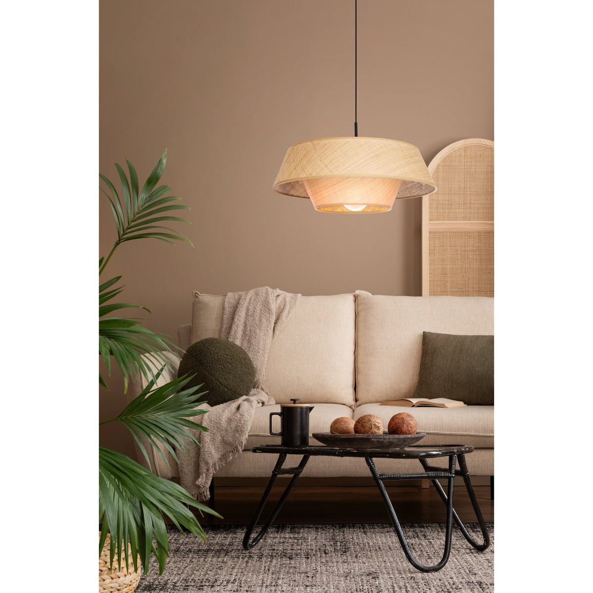 Prenvanne Pendant Light in black with a two-tier woven bast shade in natural straw tone, shown lit above a living room coffee table and beige sofa.