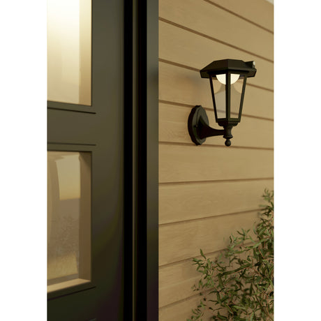 Prietta Solar Wall Light with Motion Sensor in black, a six-sided lantern-style fixture with clear glass panels and a warm white globe, mounted on wood siding beside a front door.