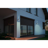 Prinorne Outdoor Wall Light in black cast aluminium with a curved half-dome hood, shown illuminated on a house exterior beside sliding doors and shutters.