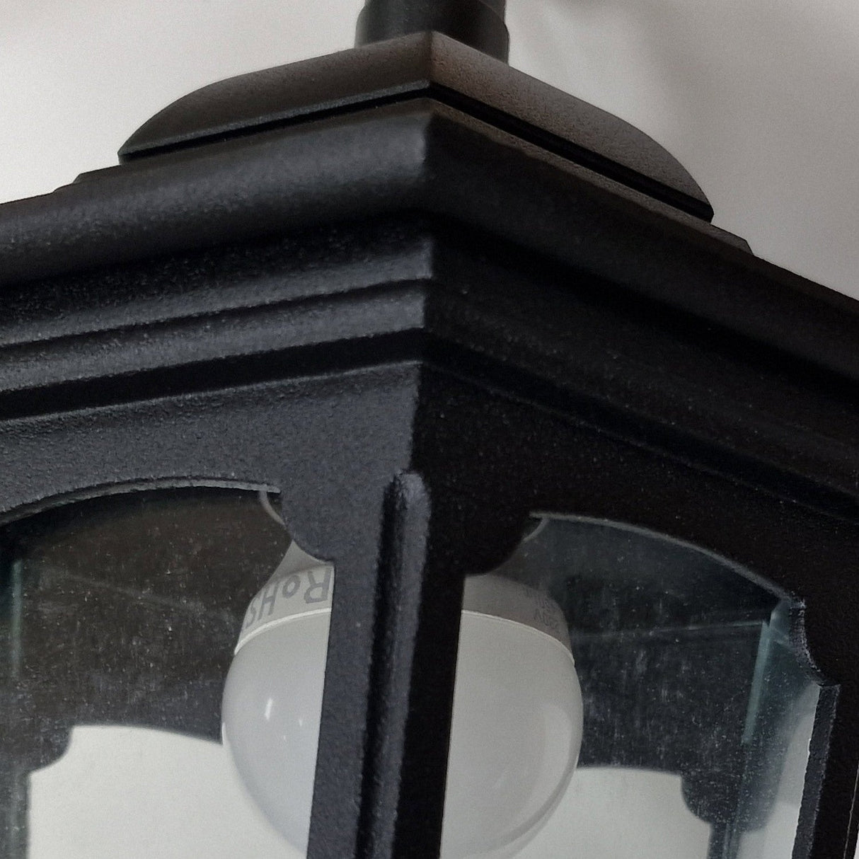 Close-up of the Parish Mini 1 Light Outdoor Chain Pendant - Black, featuring a black metal frame and frosted glass panels. The frosted bulb inside casts ambient lighting against a white wall background, with the black powder coating accentuating its sleek, modern design.