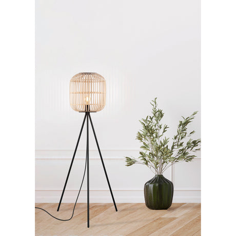 Prolbane Floor Lamp in black with a slim tripod steel base and barrel-shaped wood slat shade, shown lit in a living room beside a potted plant.