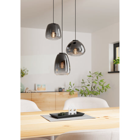 Prolbette 3 light cluster pendant in black with smoked glass shades of varying bell, dome and tapered shapes, shown hanging above a wooden dining table in a bright living space.