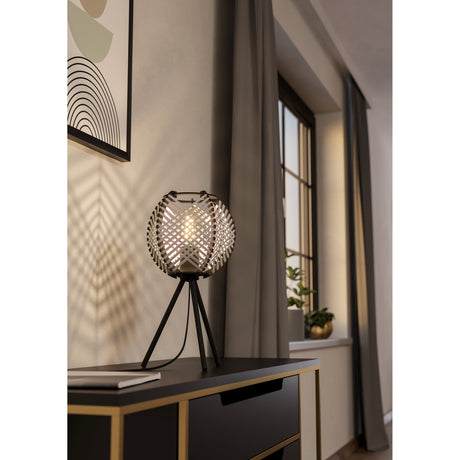 Prosena table lamp with a tripod black steel base and round woven black-and-white shade, lit on a dark sideboard beside a window and framed art.