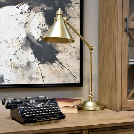 A vintage typewriter rests on a wooden desk beside the Provence 1 Light Table Lamp in aged brass, which offers adjustable illumination. Two closed books are stacked nearby. In the background, you can see an abstract painting featuring black and beige splatters.