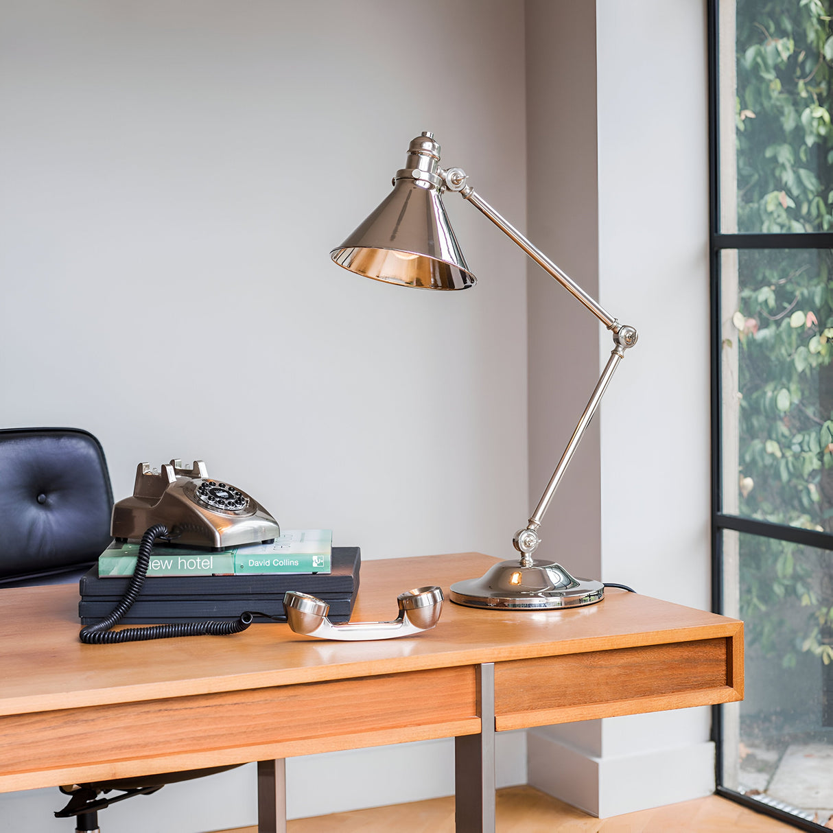 A contemporary workspace includes a wooden desk adorned with the Provence 1 Light Table Lamp in polished nickel, showcasing adjustable joints, alongside a vintage rotary telephone resting on a stack of books. A black leather chair complements the setting, all against the backdrop of a large window overlooking lush greenery.