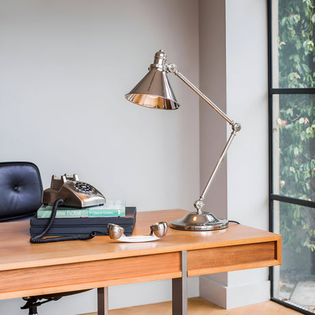 A contemporary workspace includes a wooden desk adorned with the Provence 1 Light Table Lamp in polished nickel, showcasing adjustable joints, alongside a vintage rotary telephone resting on a stack of books. A black leather chair complements the setting, all against the backdrop of a large window overlooking lush greenery.