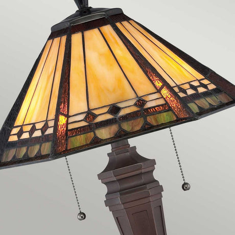 The Arden 2 Light Table Lamp - Bronze Patina features a stained glass shade with geometric patterns in warm yellow and brown tones. Its ornate base, highlighted with a bronze patina, includes two pull chains. When illuminated, the lamp casts a soft glow ideal for any room.