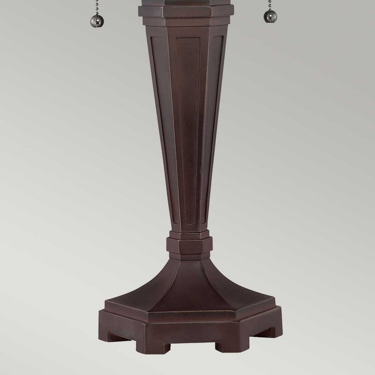 A close-up of the Arden 2 Light Table Lamp - Bronze Patina base showcases its tall, brown body with a geometric design against a flat, grey background. The base features a broad-to-narrow taper and is adorned with two metal pull chains on either side, perfectly designed to complement a stained glass shade.