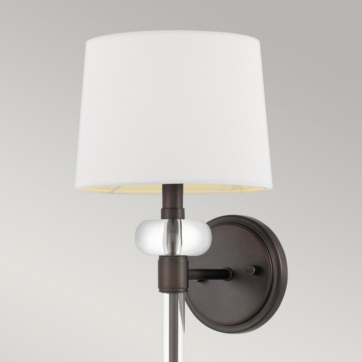 The Barbour 1 Light Wall Light in Bronze boasts a modern wall sconce design, complete with a white lampshade and Harbor Bronze finish. It features a round backplate, sleek cylindrical arm, and an elegant glass accent, embodying modern aesthetics to enhance ambient lighting in any stylish interior space.