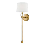 The Barbour 1 Light Wall Light - Weathered Brass, featuring a cylindrical arm, a white lampshade, and a round base, is mounted on the wall. This design seamlessly blends modern elegance with vintage allure, adding a sophisticated touch to any room.