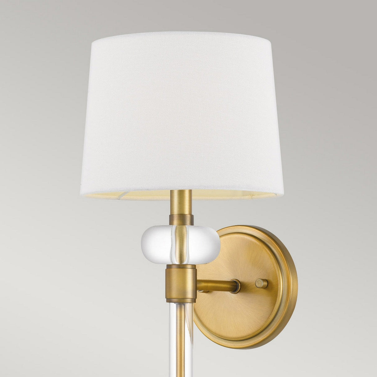 The Barbour 1 Light Wall Light in weathered brass, featuring a white fabric lampshade and a transparent decorative element hanging below it, is mounted on a circular backplate. Its vintage allure exudes modern elegance against a plain gray background.