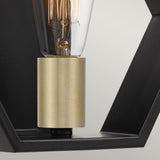 A close-up image features the Bismarck 1 Light Wall Light in black, highlighting its geometric profile. This elegant fixture includes a vintage-style Edison bulb partially encased in a black metal frame, complemented by a brass-colored socket with gold accents for added contrast. The intricately visible filament enhances the sophisticated design.