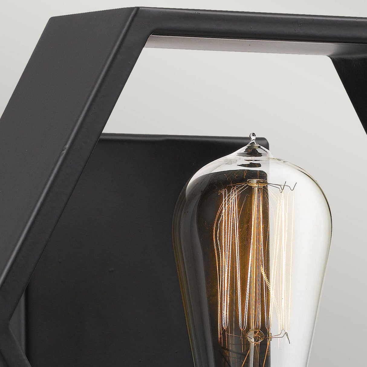 A close-up of the Bismarck 1 Light Wall Light - Black features a vintage-style exposed filament bulb encased within a modern, angular black metal frame with gold accents. The bulb casts a warm glow, accentuating the geometric profile of the fixture against a pale green background.