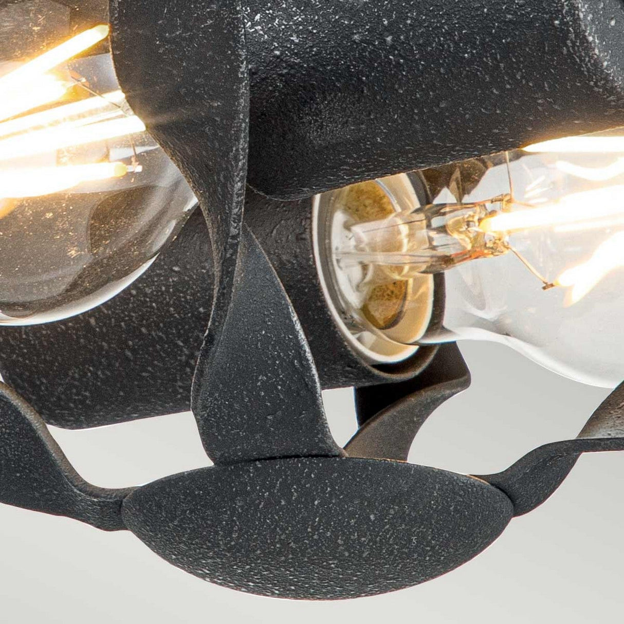 A detailed view of the Bradbury 2 Light Flush in Grey Ash, showcasing its contemporary design with a black, textured metal fixture and exposed bulbs. The intricately overlapping metal pattern encircles the bulbs, casting a warm glow reminiscent of grey ash.