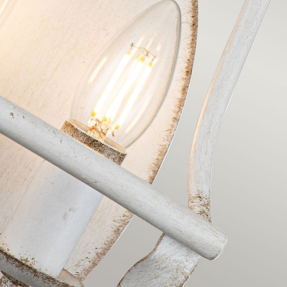 A close-up of the Bradbury 2 Light Wall Light in Antique White showcases its vintage-style bulb, which is illuminated and casting a warm glow. The sconce's worn finish enhances its antique charm against a light, neutral background that complements the cozy ambiance.