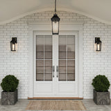 White double front doors with glass panels and silver handles are accented by a Buckley 1 Light Coastal Small Outdoor Wall Lights Lantern in black and potted shrubs in square planters. A brown doormat sits on the brick porch below a stylish hanging lantern above.