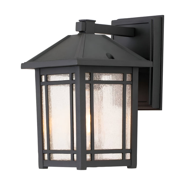 The Cedar Point Outdoor 1 Light Small Wall Lantern - Black features a vintage design with a rectangular metal frame accented by seedy glass panels. Ideal for rustic outdoor lighting, it provides a warm glow and is mounted on a square base with a dark, rustic finish.
