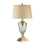 The Dennison 1 Light Table Lamp in brushed brass showcases a clear glass base with vertical grooves and an elegant square pedestal. It features a slightly tapered beige lampshade, creating an exquisite design that adds traditional elegance to any home decor.