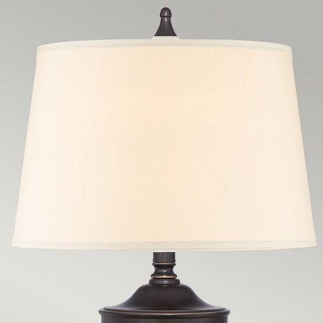 The Dennison 1 Light Table Lamp in Palladian Bronze, with its beige fabric lampshade, captures the essence of transitional interiors. Positioned against a plain gray background and currently turned off, this lamp elegantly merges classic and modern design elements.