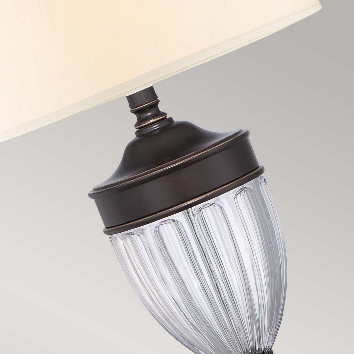 A close-up view of the Dennison 1 Light Table Lamp - Palladian Bronze reveals its elegant white lampshade and clear, textured glass body with a Palladian Bronze accent, making it ideal for transitional interiors against a softly muted gray background.