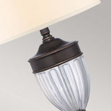 A close-up view of the Dennison 1 Light Table Lamp - Palladian Bronze reveals its elegant white lampshade and clear, textured glass body with a Palladian Bronze accent, making it ideal for transitional interiors against a softly muted gray background.