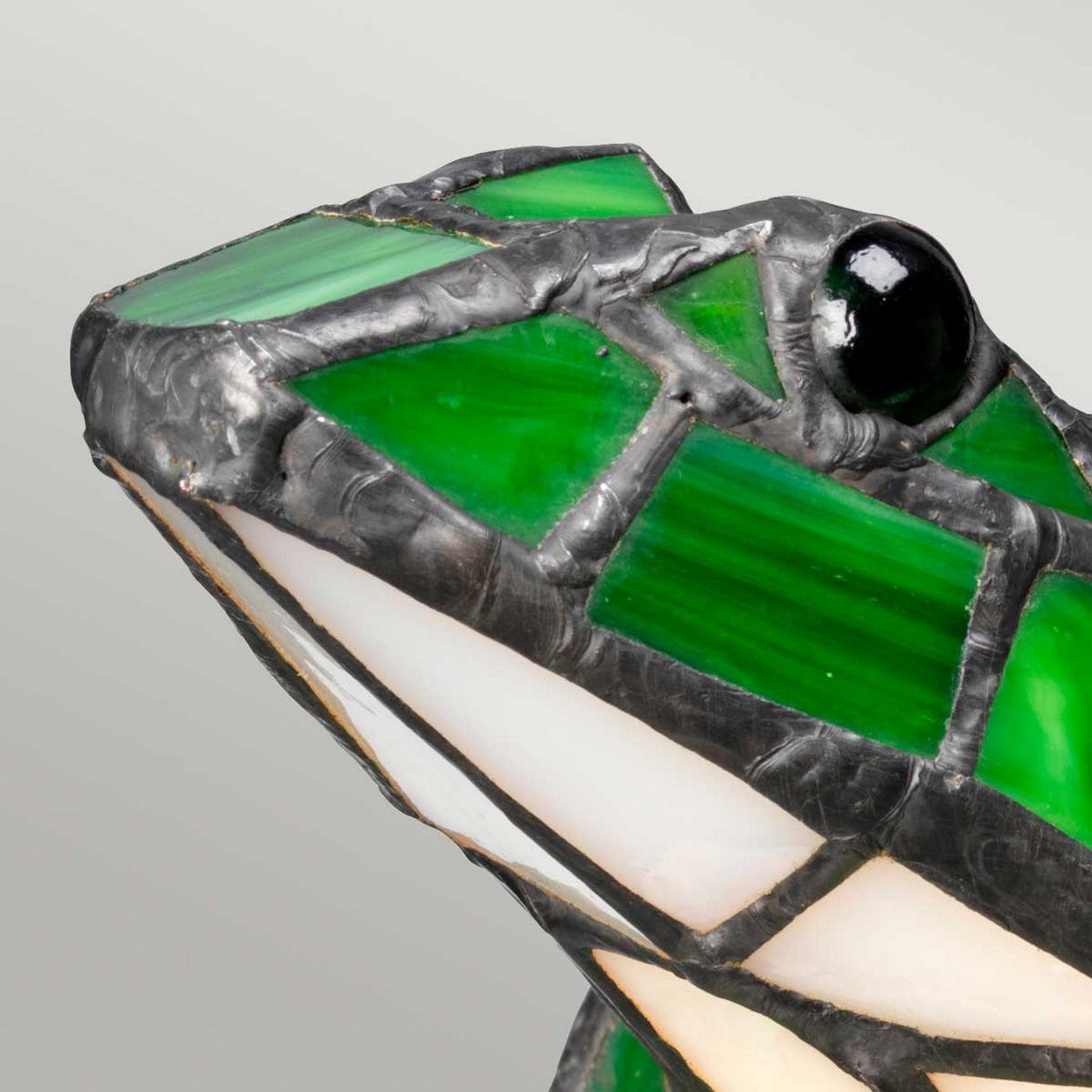 Close-up of a Tiffany Animal Lamps Frog Lamp in vintage bronze, showcasing green and white stained glass pieces with a black, shiny eye. The frog features a geometric design with visible lead came lines connecting the segments.