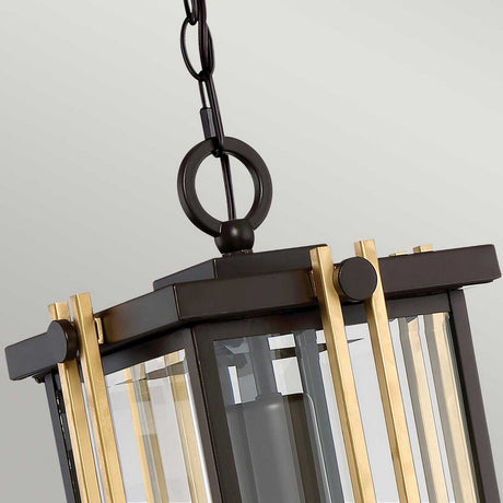 Close-up of the Goldenrod Outdoor 1 Light Medium Chain Pendant in bronze, showcasing a sleek black metal frame adorned with vertical brass accents and clear glass panels. This piece exudes mid-century lighting charm, elegantly suspended by a black chain with a circular hook at the top.