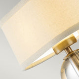 A close-up of a contemporary table lamp featuring a beige fabric lampshade and a brushed brass base, set against a neutral backdrop. Ideal for modern interiors, the gentle lighting captures the warmth reminiscent of the Gotham 1 Light Wall Light - Brushed Brass, fostering an inviting atmosphere.