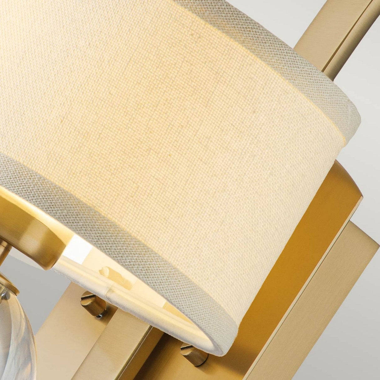 Close-up of the Gotham 1 Light Wall Light, showcasing its brushed brass body and beige fabric lampshade with a subtle texture. The warm light softly illuminates the surrounding area, making it ideal for modern interiors.