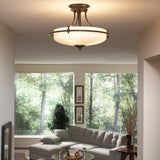 A modern living room showcases a Griffin 4 Light Semi-Flush in Palladian Bronze, equipped with an etched glass shade that brightens the area. A large window presents a lush green view over a gray sectional sofa, while two framed pictures and a vase with tall branches enhance the room's charm.
