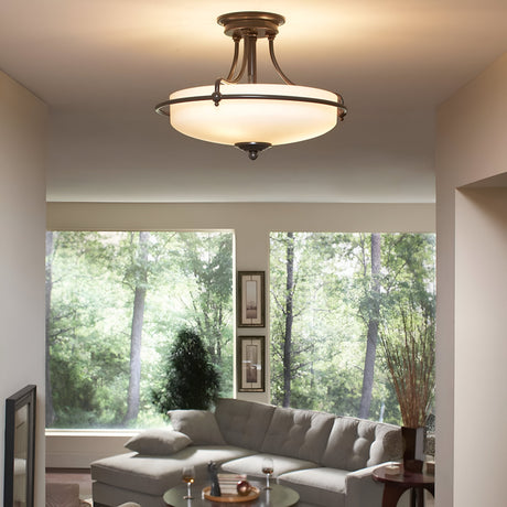 A modern living room showcases a Griffin 4 Light Semi-Flush in Palladian Bronze, equipped with an etched glass shade that brightens the area. A large window presents a lush green view over a gray sectional sofa, while two framed pictures and a vase with tall branches enhance the room's charm.