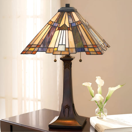 An Inglenook 2 Light Table Lamp in Valiant Bronze, featuring a Tiffany-style stained glass shade, rests on a dark wooden table. Beside it, a small glass vase contains three white calla lilies. Soft natural light filters through a sheer curtain in the background.