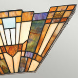 A close-up of the Inglenook 2 Light Tiffany Wall Light in Valiant Bronze showcases a stained glass panel with geometric patterns in shades of amber, brown, purple, and blue. Inspired by classic Arts & Crafts lighting, the design features elongated triangular shapes and a symmetrical arrangement, all framed by steel outlining each segment.