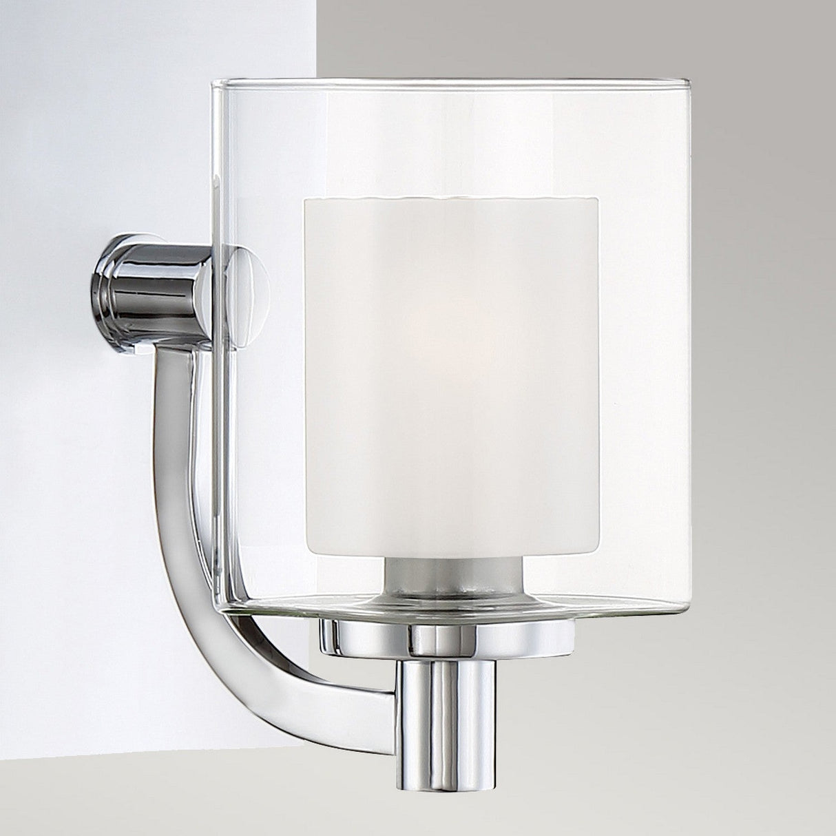 The Kolt 1 Light Wall Light in polished chrome features a cylindrical, frosted glass shade that is elegantly mounted on a light gray wall. This bathroom wall light showcases a minimalist, curved design, providing a sleek and contemporary touch to enhance any modern bathroom lighting scheme.
