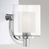 The Kolt 1 Light Wall Light in polished chrome features a cylindrical, frosted glass shade that is elegantly mounted on a light gray wall. This bathroom wall light showcases a minimalist, curved design, providing a sleek and contemporary touch to enhance any modern bathroom lighting scheme.