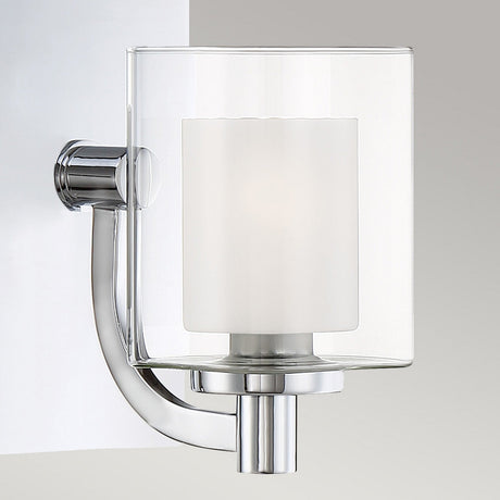 The Kolt 1 Light Wall Light in polished chrome features a cylindrical, frosted glass shade that is elegantly mounted on a light gray wall. This bathroom wall light showcases a minimalist, curved design, providing a sleek and contemporary touch to enhance any modern bathroom lighting scheme.