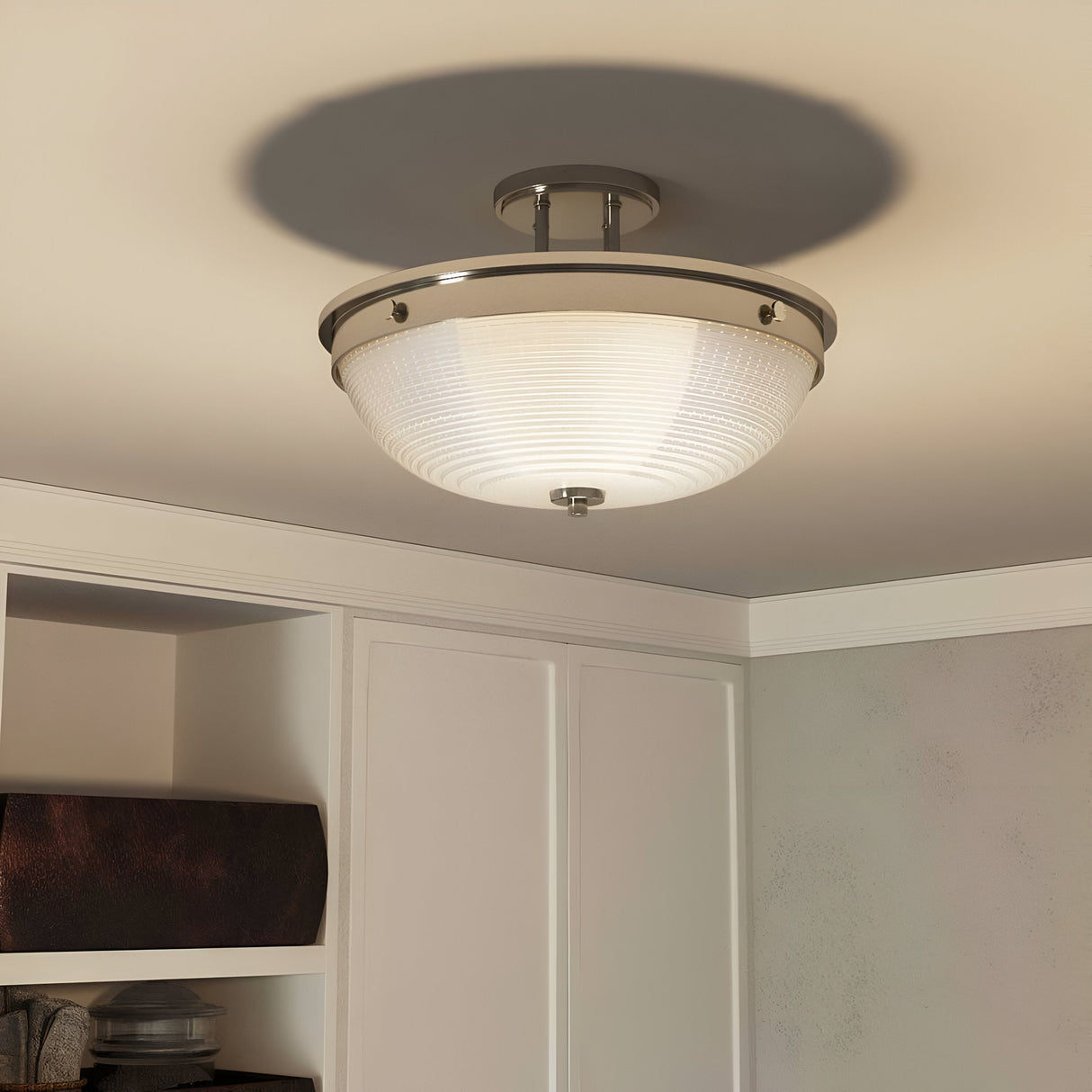 The Mantle 2 Light Semi-Flush - Imperial Silver ceiling fixture showcases a contemporary design with its circular frosted glass and Imperial Silver metallic rim. When installed on a plain white ceiling in a room featuring white cabinets and shelves, this fixture emits a soft glow and creates subtle shadows, enriching the elegance of the space.