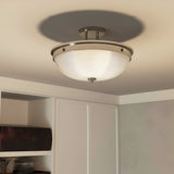 The Mantle 2 Light Semi-Flush - Imperial Silver ceiling fixture showcases a contemporary design with its circular frosted glass and Imperial Silver metallic rim. When installed on a plain white ceiling in a room featuring white cabinets and shelves, this fixture emits a soft glow and creates subtle shadows, enriching the elegance of the space.