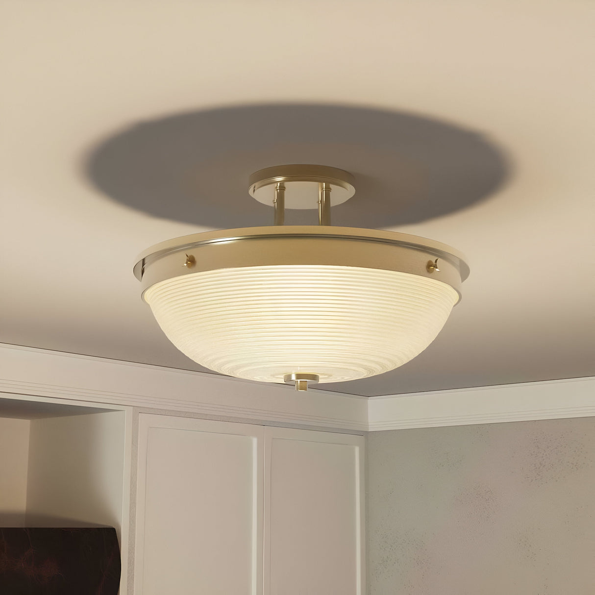 Close-up of the Mantle 2 Light Semi-Flush in weathered brass, featuring prismatic glass and a ribbed, frosted shade that casts a soft glow. Neutral-colored walls surround the space, offering a glimpse of a cabinet below the softly illuminated fixture.