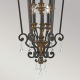 The Marquette-B 6 Light Chandelier - Bronze is an elegant fixture showcased by its black and gold metal finish, intricate scrollwork, and hanging crystal accents. This bronze chandelier features six candle-style lights adorned with sparkling crystal drops.