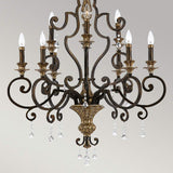 The Marquette 9 Light Chandelier in bronze combines vintage style with ornate dark metal scrollwork and features nine candle-style lights adorned with elegant crystal drops hanging from its arms. When illuminated, it casts a warm and inviting glow against a neutral background.