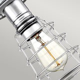 A close-up of the Mixon 1 Light Semi-Flush in polished chrome showcases a vintage-style lightbulb encased in a silver metal cage, emitting a warm glow. This fixture enhances the industrial-chic design against a plain background.