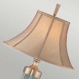 A close-up of the Monteverde 1 Light Table Lamp - Green/Aged Brass, highlighting its decorative design with a beige textured lampshade and an aged brass finish finial. The brass-colored base beautifully complements its glass body. The lampshade tilts slightly to the right against a neutral background.