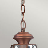 A close-up showcases the Newbury Outdoor 1 Light Medium Chain Pendant in a lacquered aged copper finish. Its chain and wiring are visible above the decorative top, enhanced by clear glass panels. The fixture contrasts beautifully against a plain light grey background, highlighting its charm and IP23-rated durability.