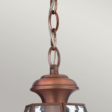 A close-up showcases the Newbury Outdoor 1 Light Medium Chain Pendant in a lacquered aged copper finish. Its chain and wiring are visible above the decorative top, enhanced by clear glass panels. The fixture contrasts beautifully against a plain light grey background, highlighting its charm and IP23-rated durability.