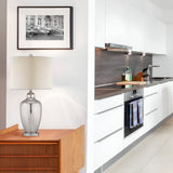 A modern kitchen with white cabinets and stainless steel appliances is adjacent to a hallway featuring a wooden drawer adorned with the Nicolls 1 Light Table Lamp in Mercury Glass. A framed black-and-white photo hangs above the drawer, adding charm to the bright, minimalist space.