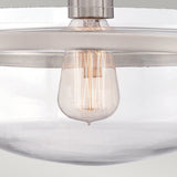 A close-up image showcases the Penn Station 1 Light Semi-Flush in Brushed Nickel, featuring a vintage-style Edison bulb in a clear glass light fixture that exudes industrial charm. The visible glowing filament is beautifully complemented by brushed nickel fixtures securing the glass. The background is a neutral color.