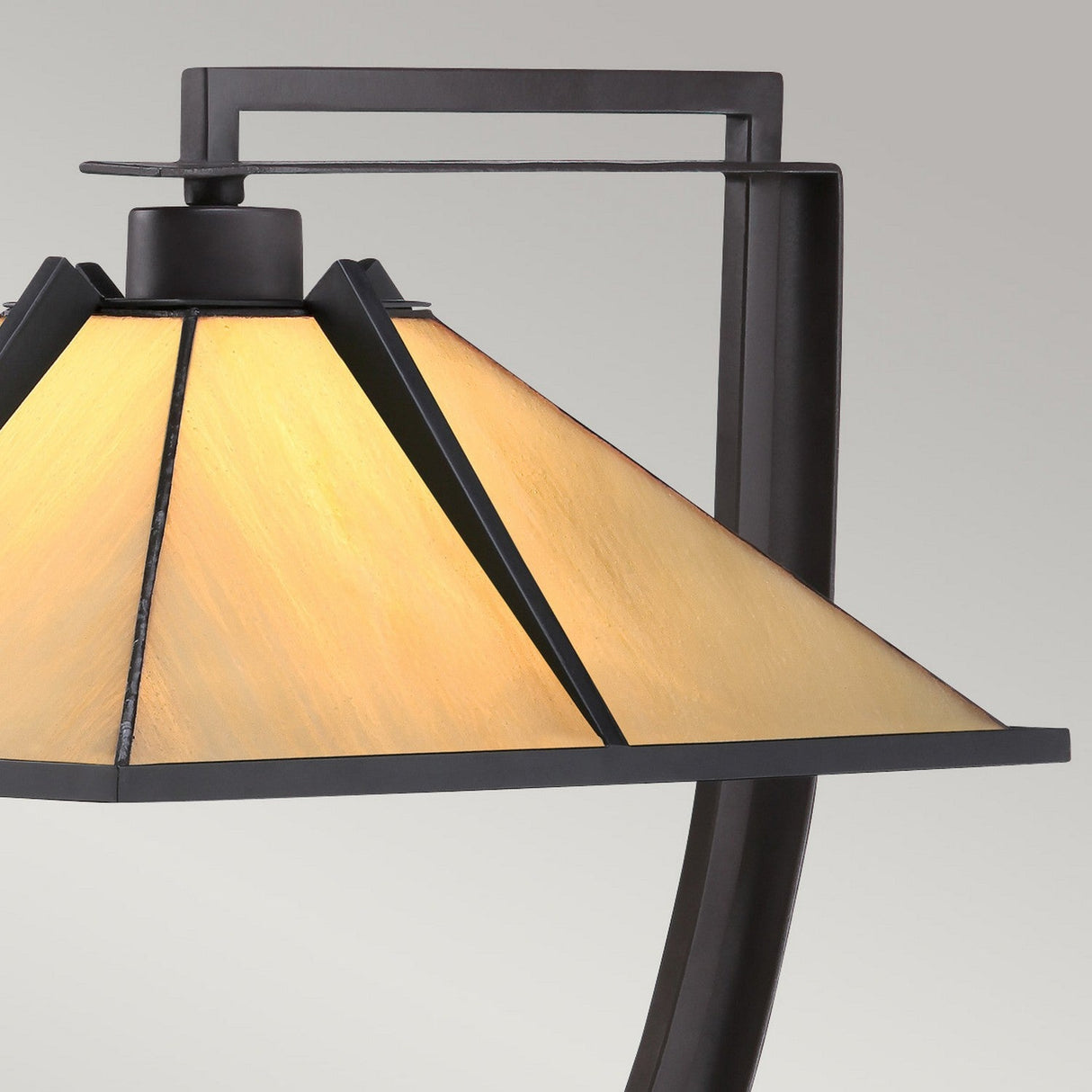 Close-up of the Pomeroy 1 Light Tiffany Table Lamp featuring a Western Bronze finish, showcasing a hand-crafted pyramid-shaped glass shade that emits a warm, ambient glow. The backdrop is a neutral gray color.