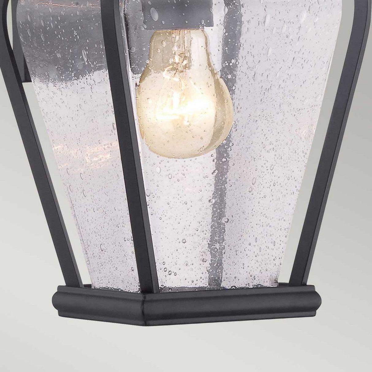 A close-up of the Province 1 Light Wall Lantern in black, featuring seeded glass panels. Crafted from durable cast aluminum, the fixture houses a glowing light bulb within its black metal framework, creating a warm glow.